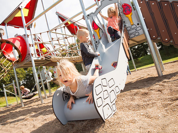Pige rutsjer ned af rutsjebane på stort legesystem som har legetårne med hængebro.